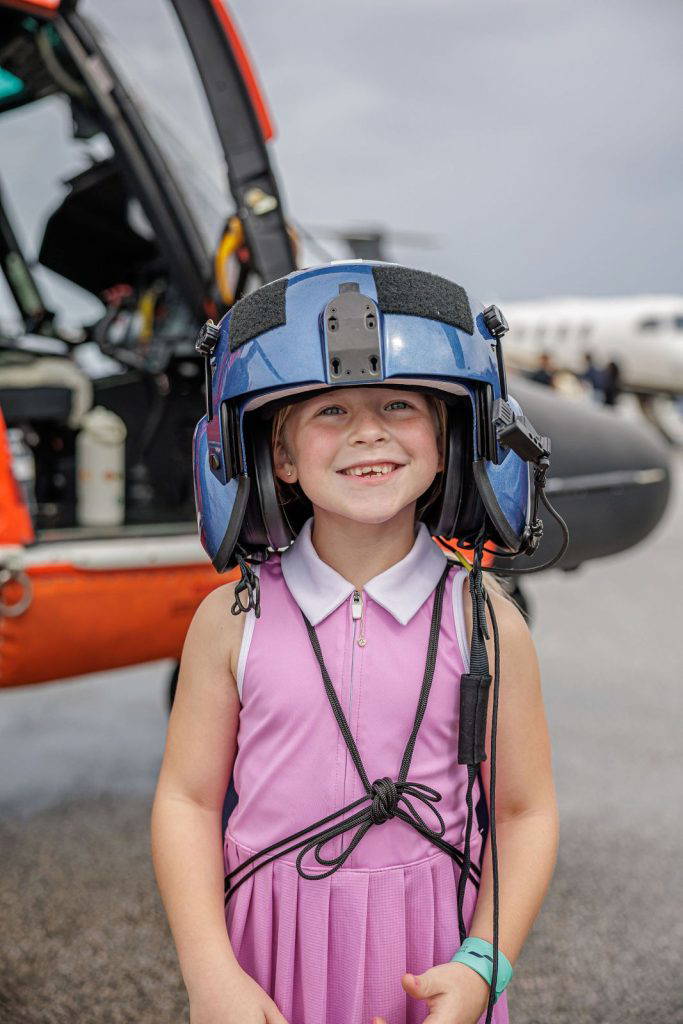 A girl grins in a big flight helmet next to the Coast Guard helicopter