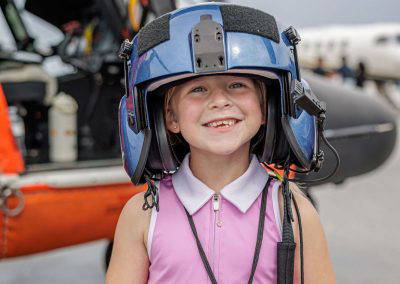 A girl smiles in a flight helmet in front of the helicopter