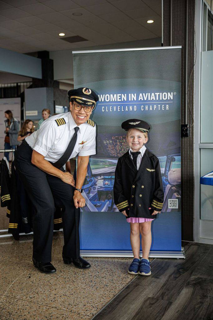 A smiling pilot poses with a little girl dressed in a pilot uniform beside the Women in Aviation banner