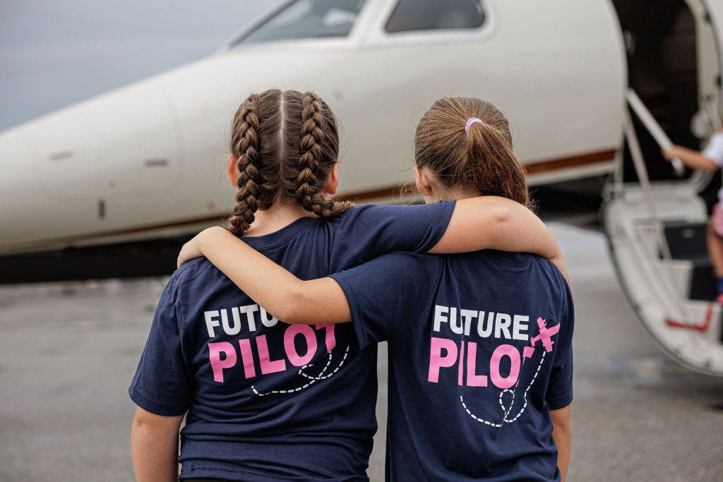 Two girls in Future Pilot shirts, arm in arm beside a jet